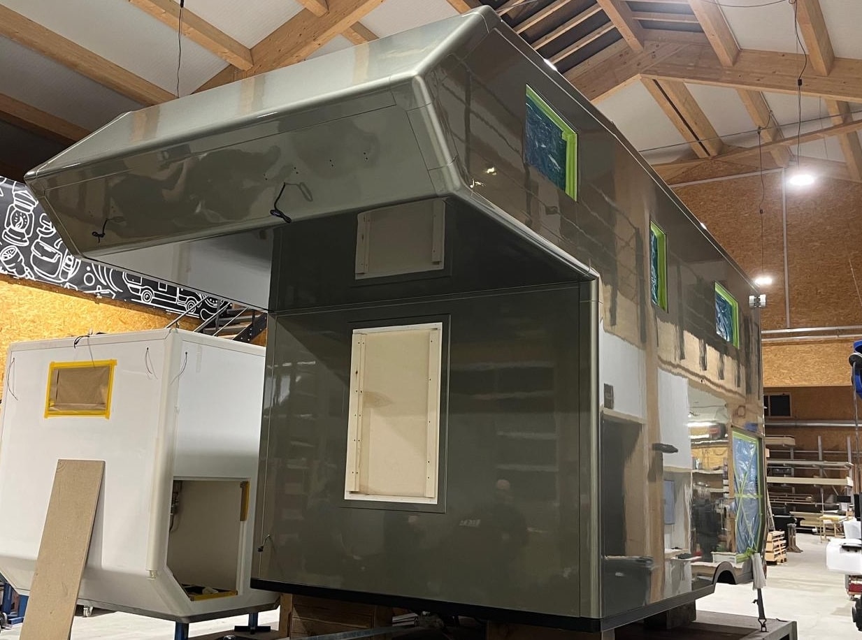 Grünes Wohncontainer-Modul in einer Werkhalle, mit Fenstern, Türen und Holzdecken, daneben weitere Bauteile.